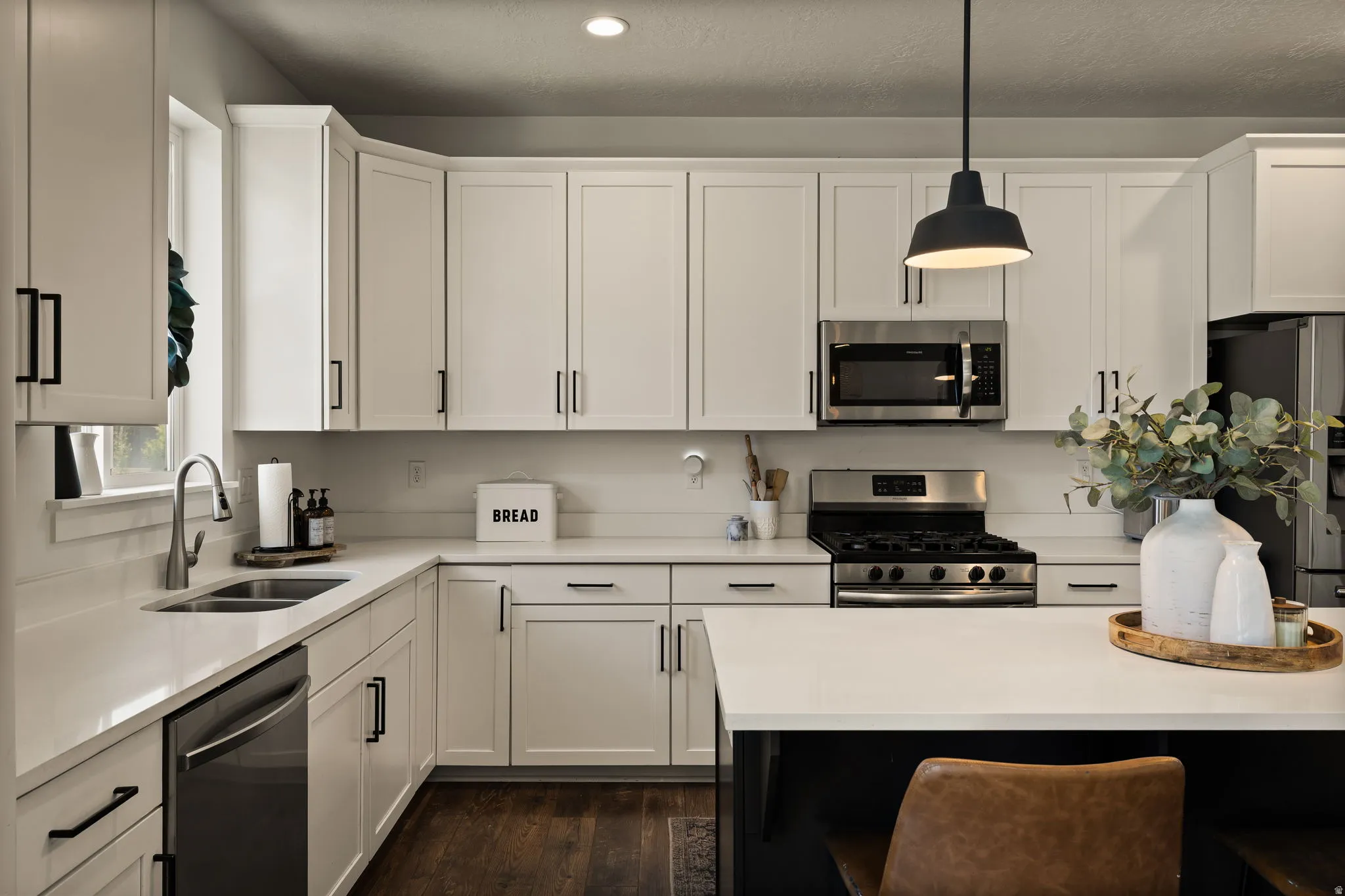 Modern kitchen featuring white shaker cabinetry, stainless steel appliances including a gas range and microwave, light-toned countertops, and dark wood-finish flooring