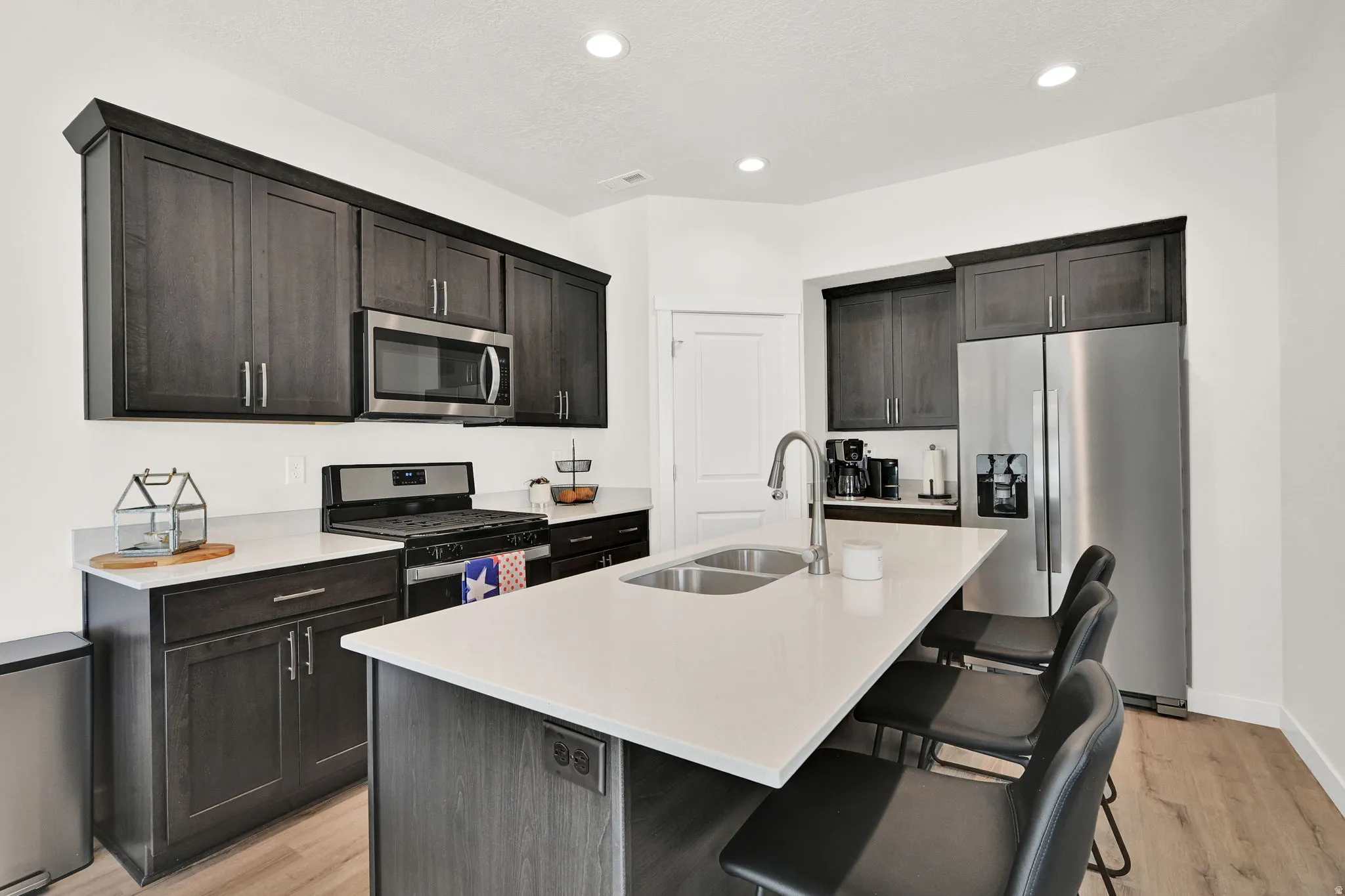 Contemporary kitchen featuring dark wood cabinetry, stainless steel appliances, a center island with double sink, light-toned countertops, and recessed lighting