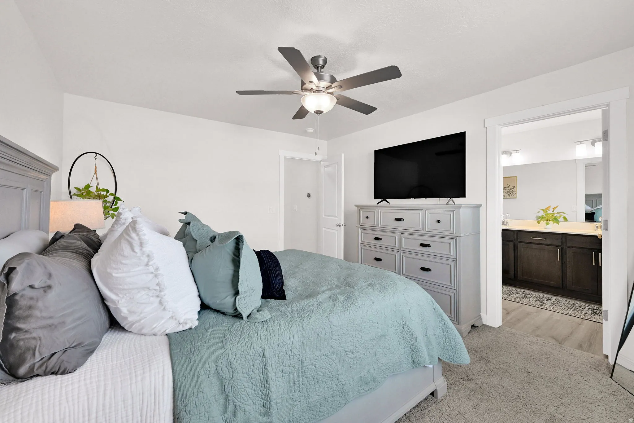 Carpeted room featuring a ceiling fan with integrated lighting, white walls, and a substantial doorway revealing a bathroom with a dual vanity, light-colored countertop, and dark wood-finish cabinetry