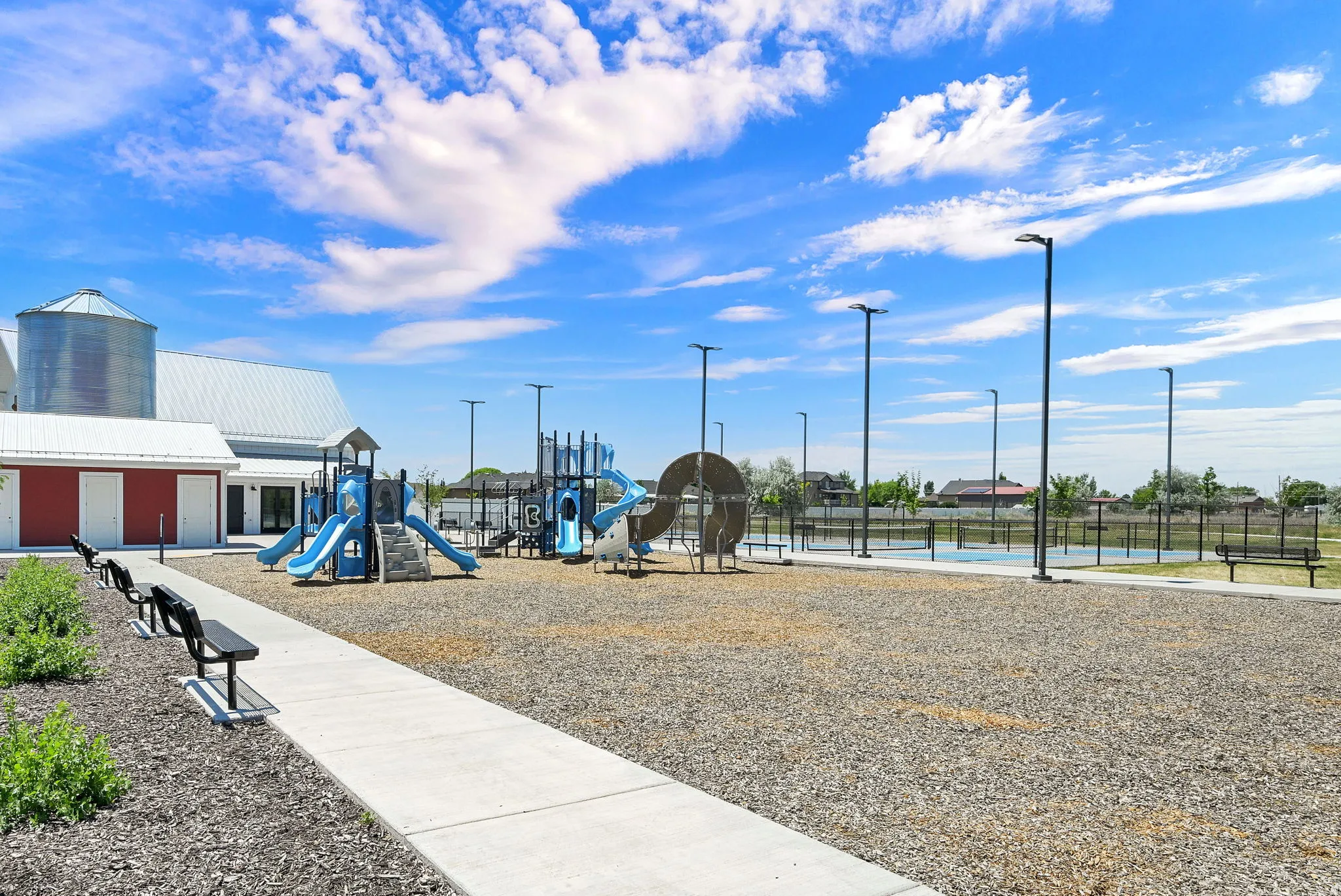 Community recreation area featuring multiple playgrounds, hardscaping, a barn-style structure with a silo, modern light posts, and paved walking paths