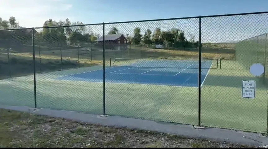 Fenced tennis court featuring a blue playing surface and light green surrounds, with a black chain-link fence