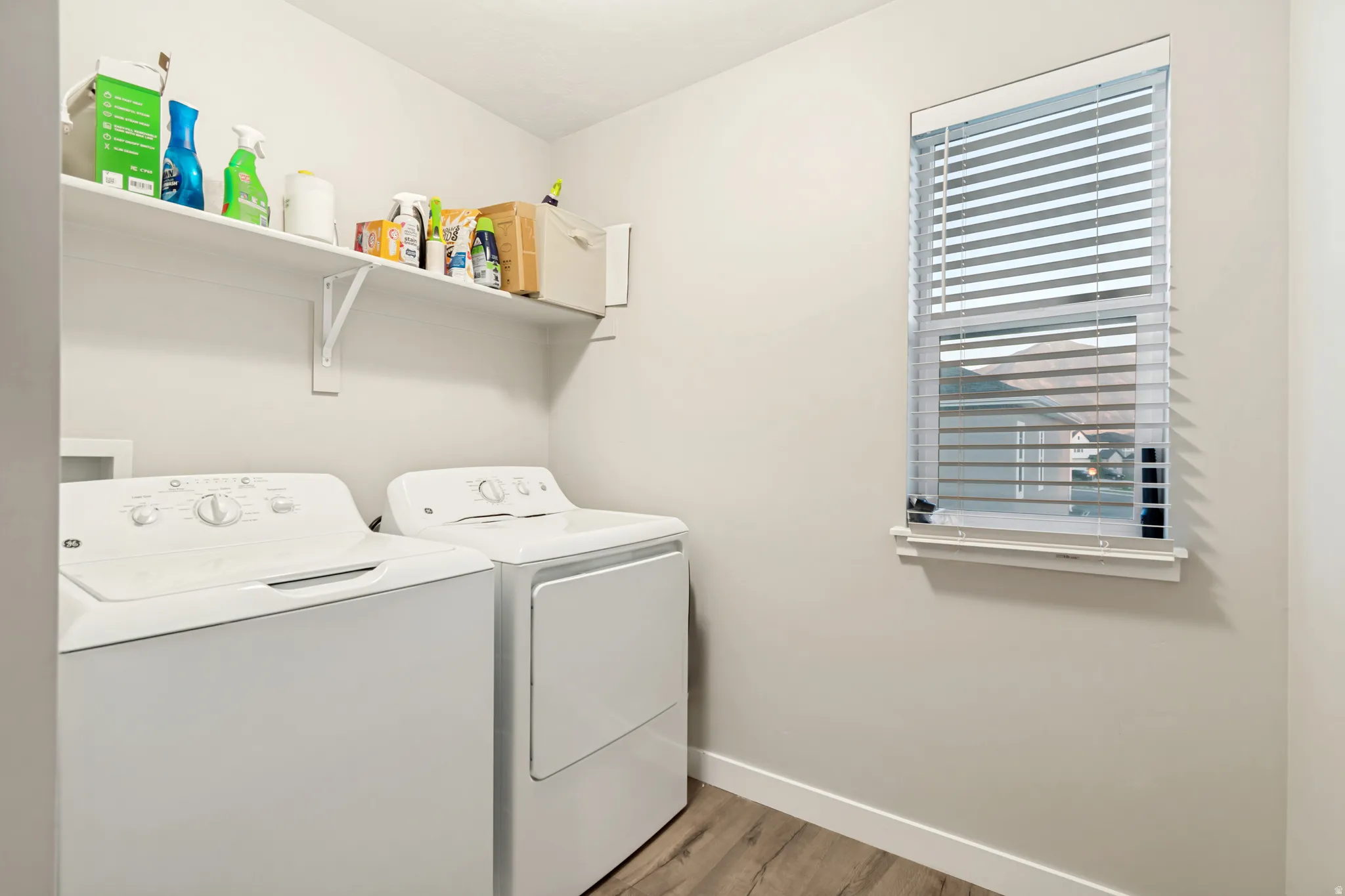 Dedicated laundry area featuring a white shelving unit, wood-finish flooring, and a window with horizontal blinds