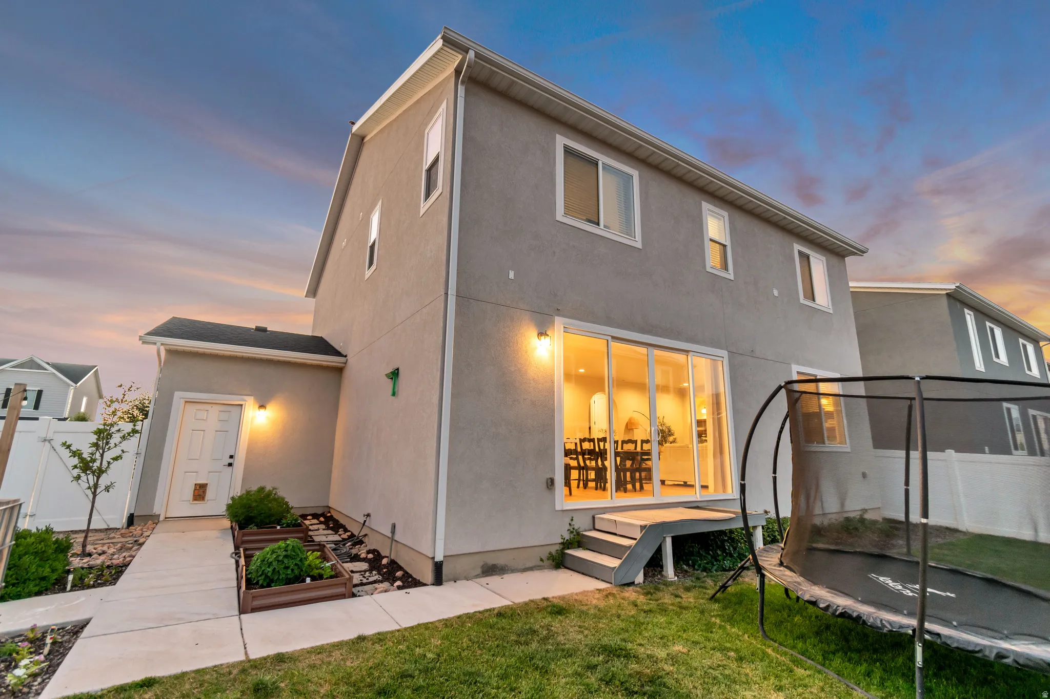 Rear exterior of a stucco home featuring a concrete patio, raised garden beds, and a white privacy fence