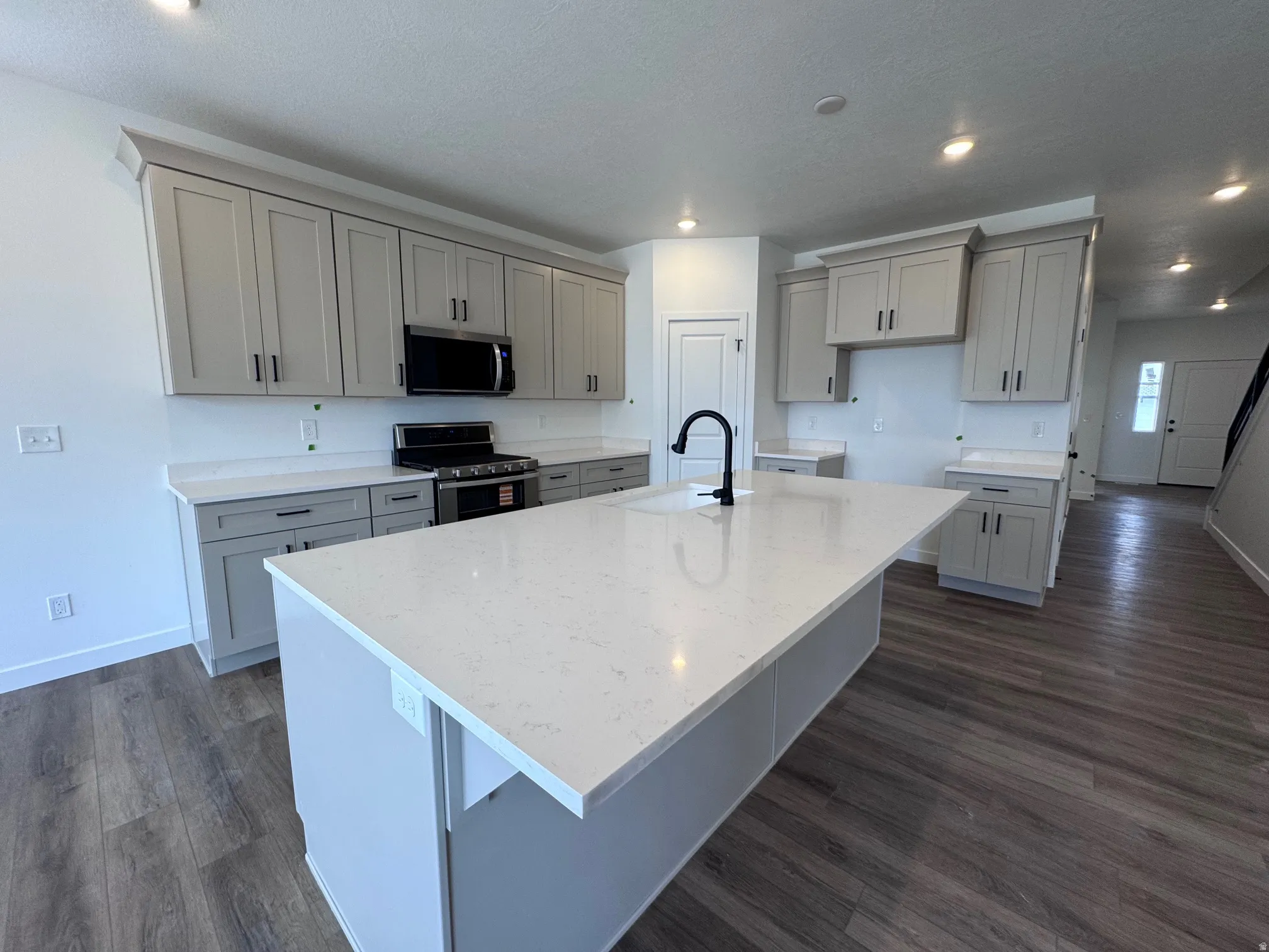 Contemporary kitchen featuring an oversized island with a white countertop and integrated sink, light gray cabinetry with black hardware, a black gas range, a built-in microwave, and wood-finish flooring