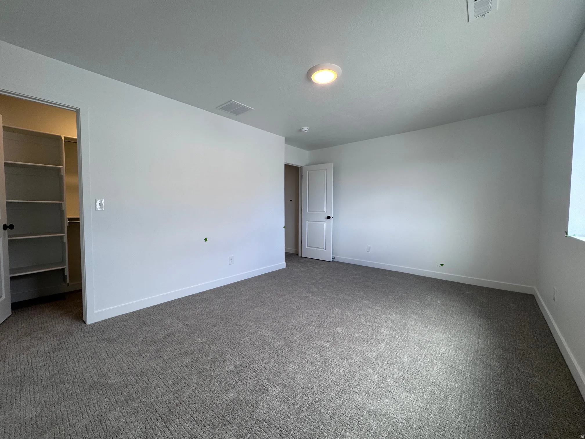 Spacious room featuring neutral carpeting, white painted walls, a built-in closet with shelving, a white door with black hardware, and a ceiling-mounted light fixture
