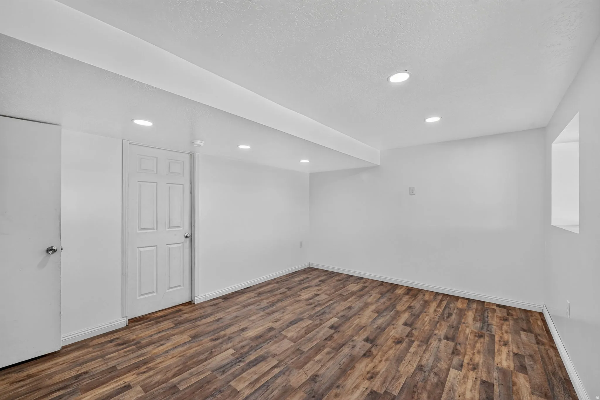 Spacious room featuring wood-finish flooring, white painted walls, recessed lighting, and a white paneled door