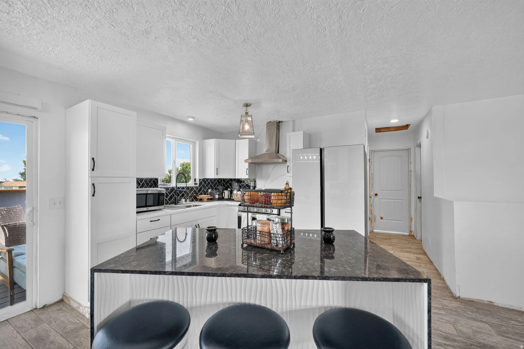 Modern kitchen featuring an island with a dark countertop and textured white paneling, white cabinetry with black hardware, a stainless steel range hood, a black and white geometric tile backsplash, and wood-finish flooring