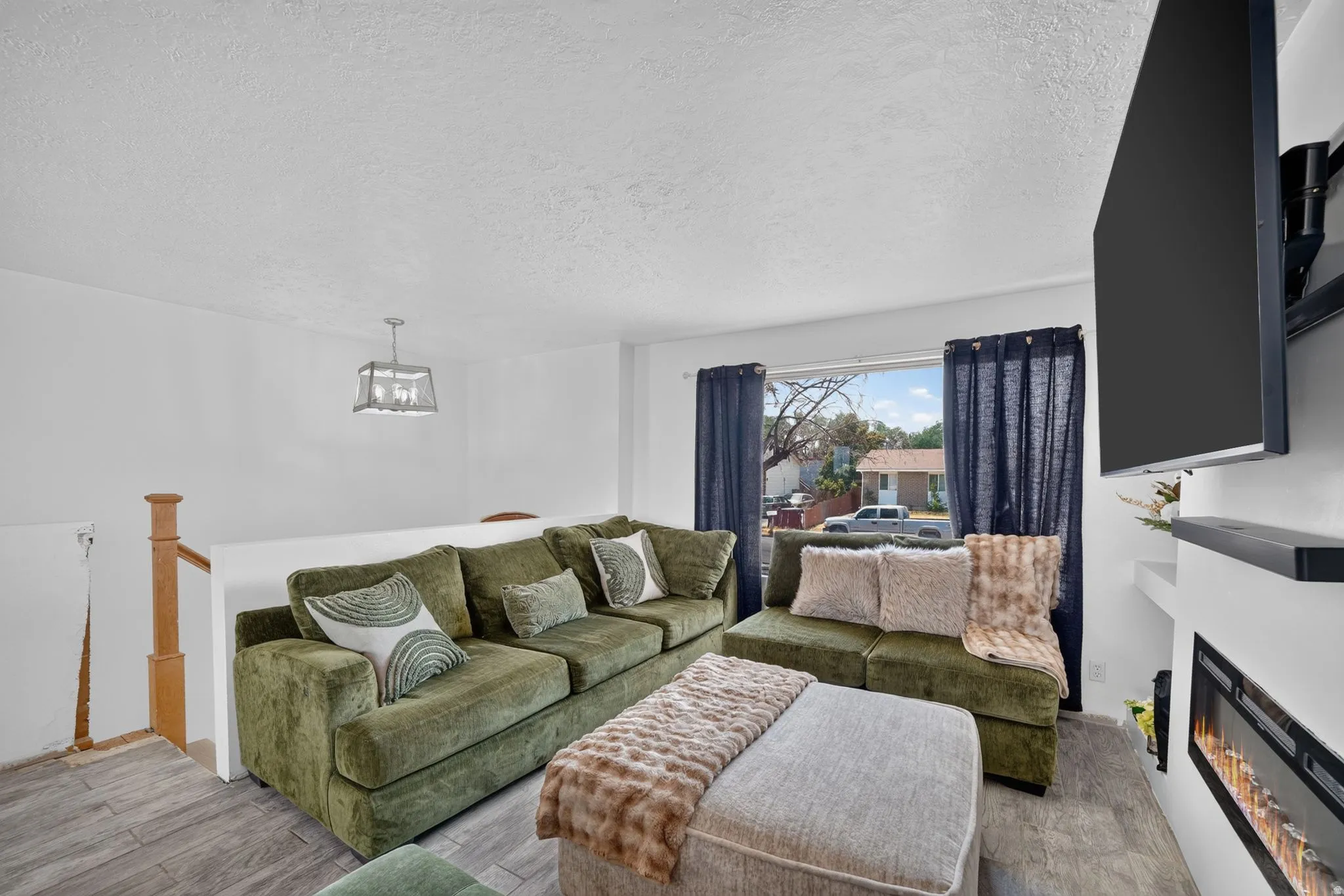 Living area featuring wood-finish flooring, a contemporary ceiling fixture, a large window with blue drapery, and a built-in electric fireplace with an integrated mantel