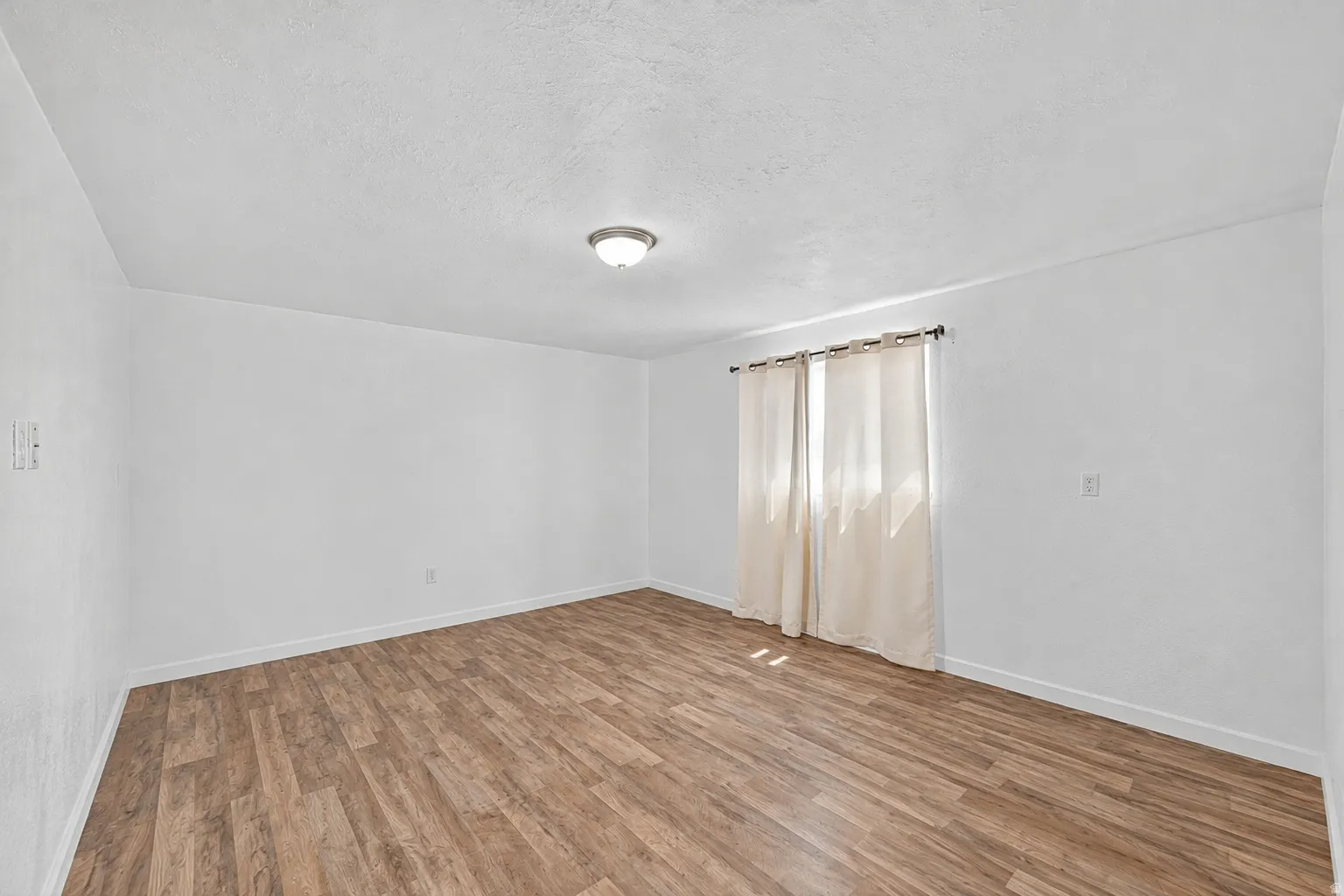 Spacious room with wood-finish flooring, white walls, and a window with light-colored drapes