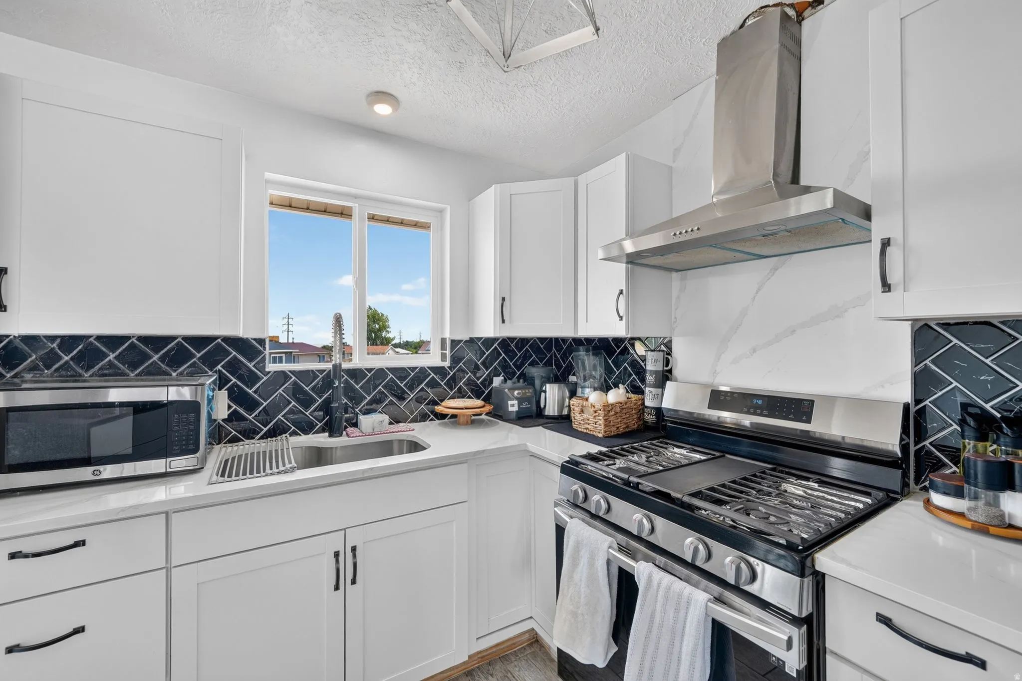 Modern kitchen featuring white shaker cabinetry, white countertops, a black herringbone tile backsplash, a stainless steel range with a coordinating hood, and a stainless steel undermount sink with a gooseneck faucet