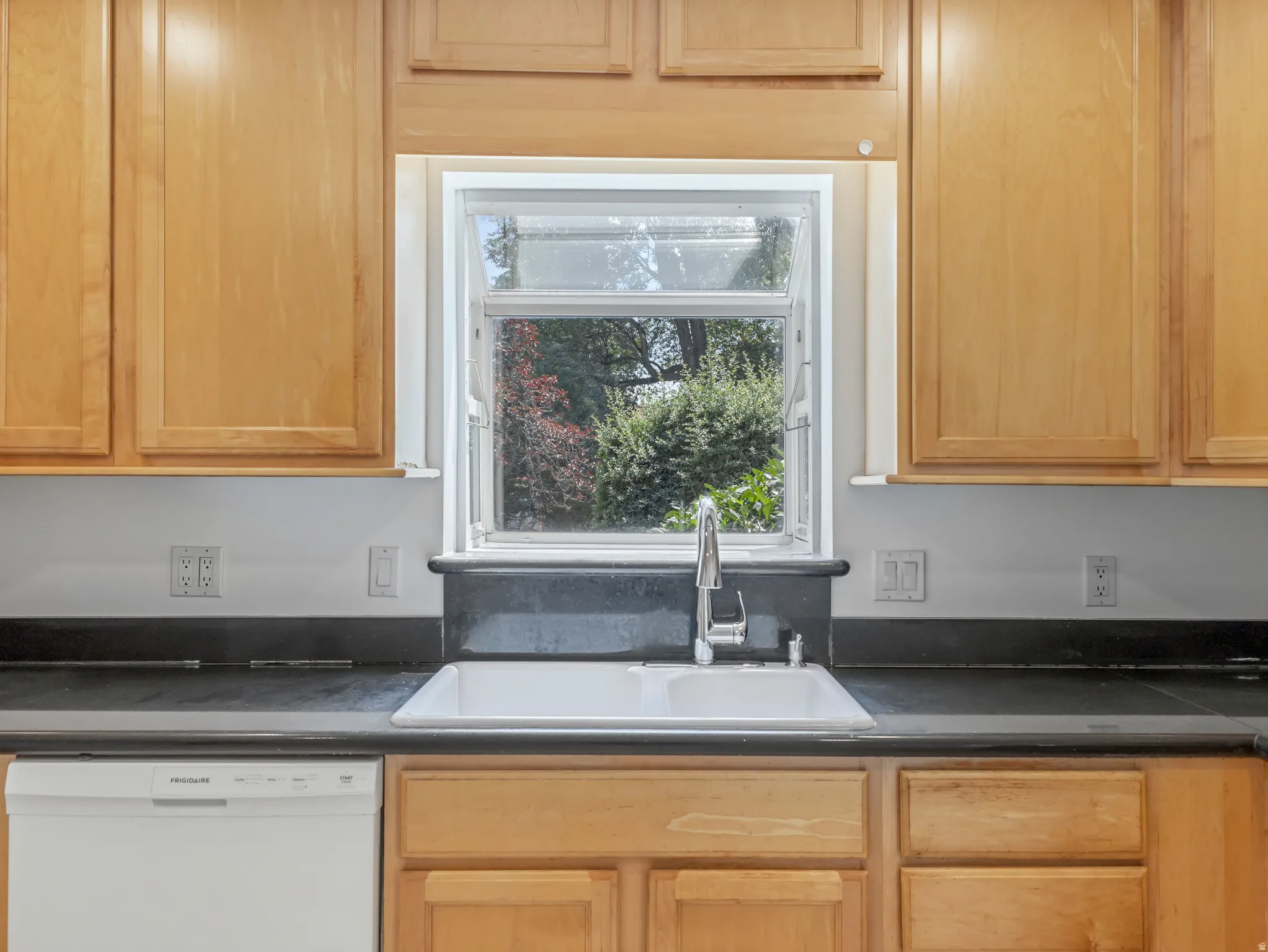 Kitchen sink area featuring light wood cabinetry, white wall paint, dark countertops, a white double basin sink, and a stainless steel faucet