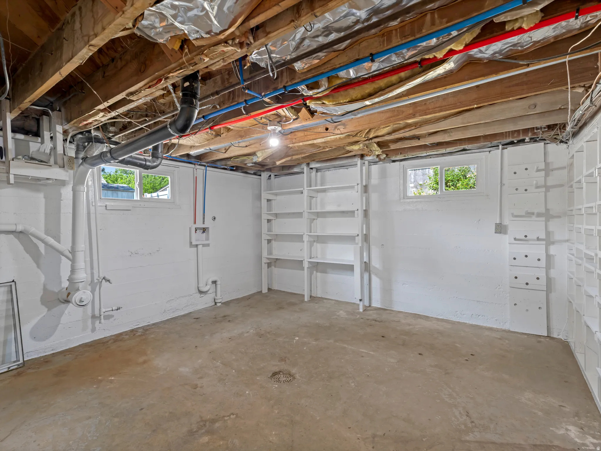 Unfinished basement space featuring exposed ceiling joists, two windows providing natural light, and built-in shelving units