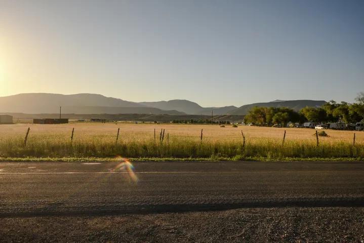 Paved road frontage with a rustic fence bordering a golden field, set against a backdrop of distant mountains