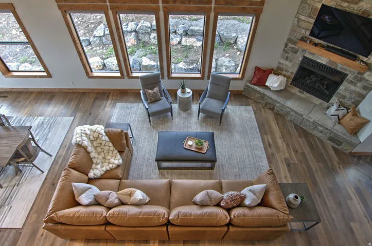 Open-concept living space featuring wood-finish flooring, a stone-clad fireplace with a hearth, and multiple large windows with wood trim
