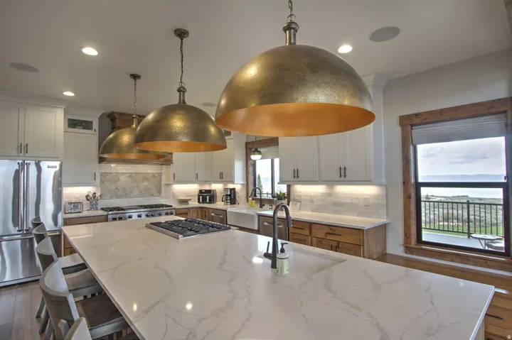 Gourmet kitchen featuring a large island with a cooktop, three distinctive pendant lights, white cabinetry, integrated stainless steel appliances, and a window with a distant landscape
