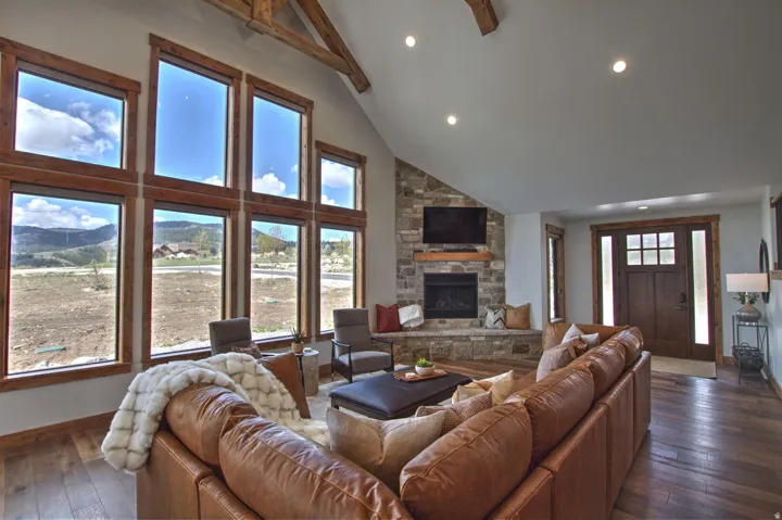 Vaulted living room featuring a stone-clad fireplace with a hearth, wood-finish flooring, exposed ceiling beams, recessed lighting, and a prominent wall of windows with wood trim
