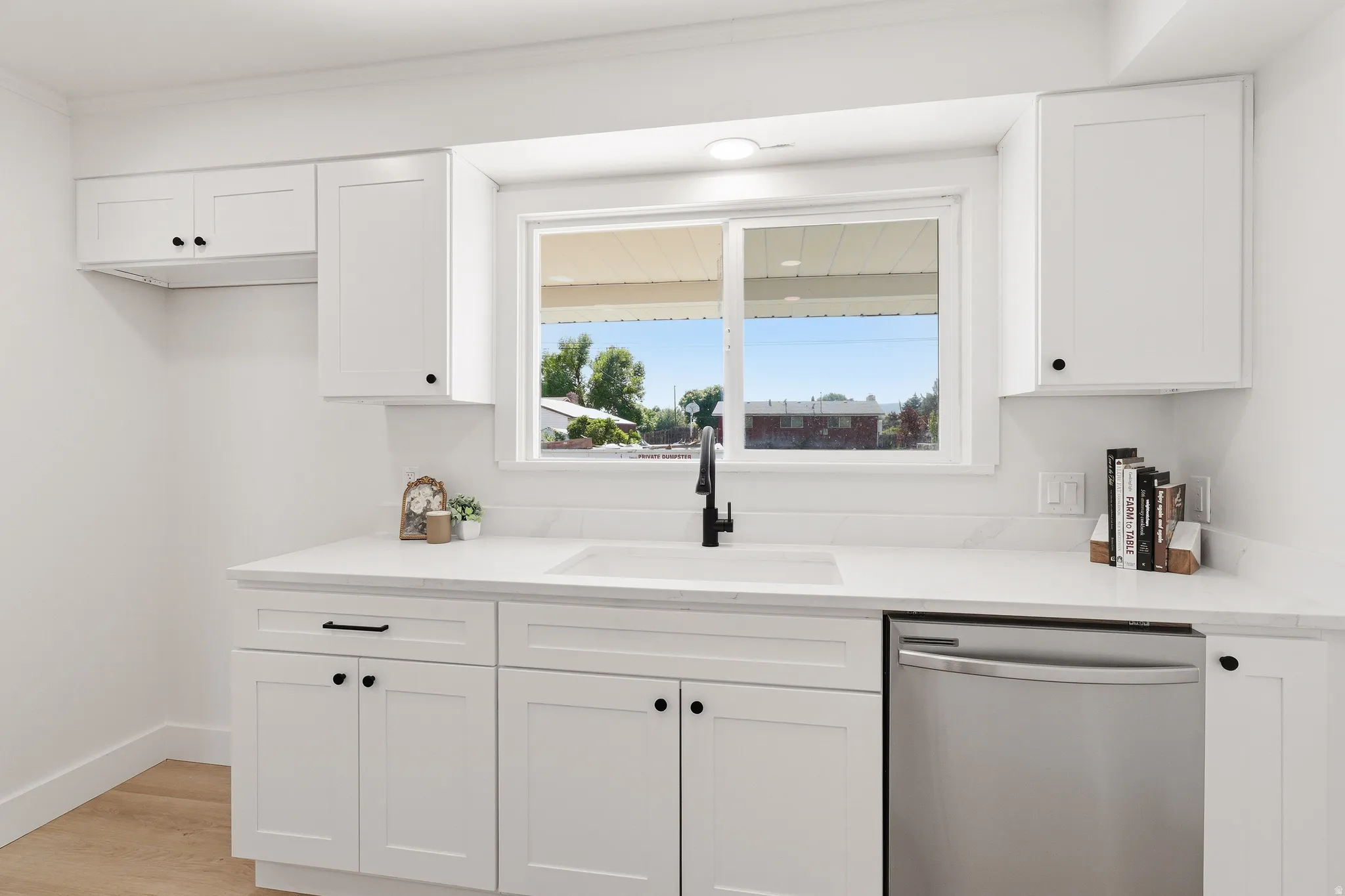 Modern kitchen featuring white shaker cabinetry, white countertops, a stainless steel dishwasher, a single basin sink with a matte black faucet, and light wood-finish flooring