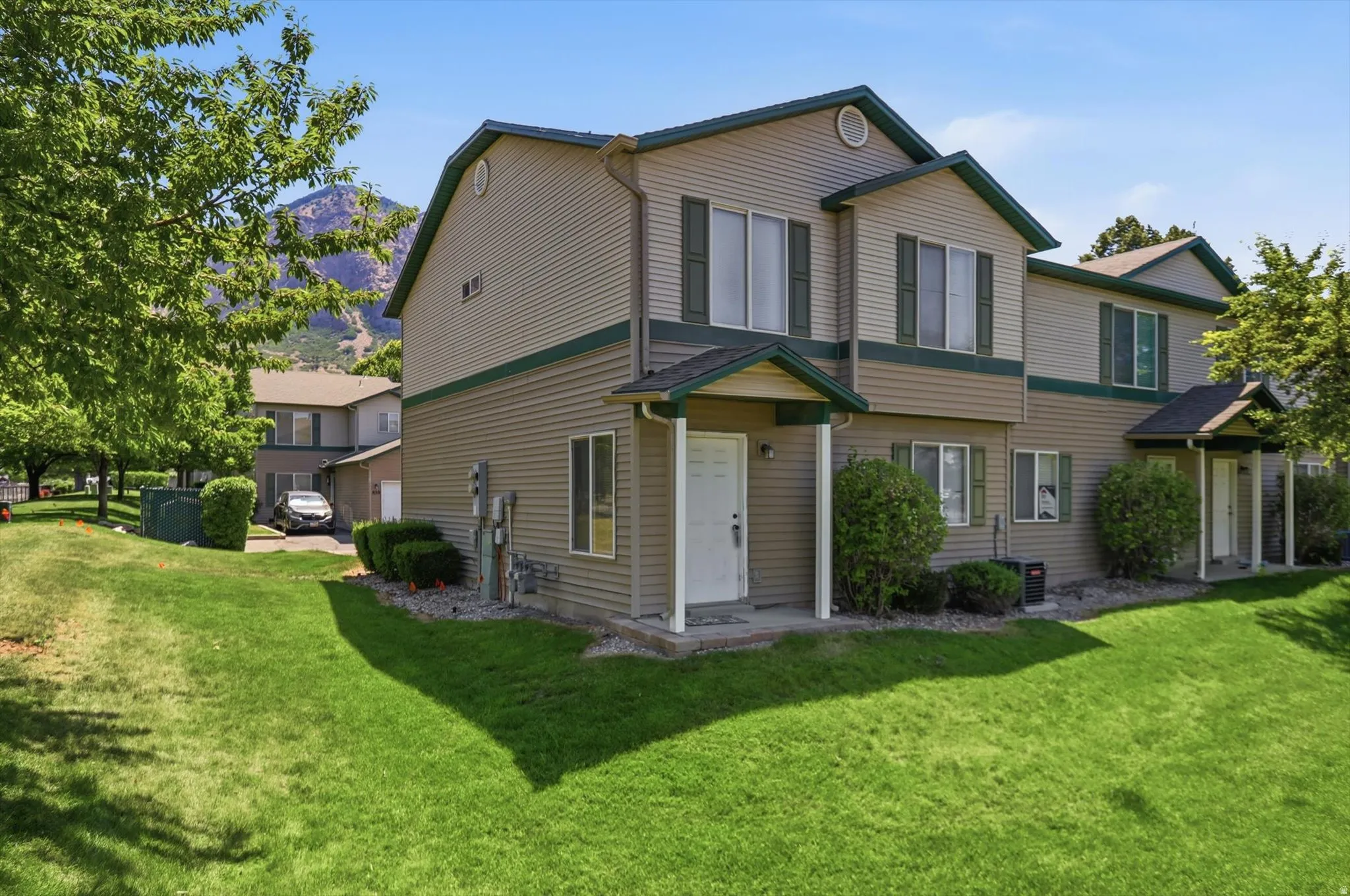 Two-story townhouse with beige siding and dark green trim, featuring a covered front entry and a mountain backdrop