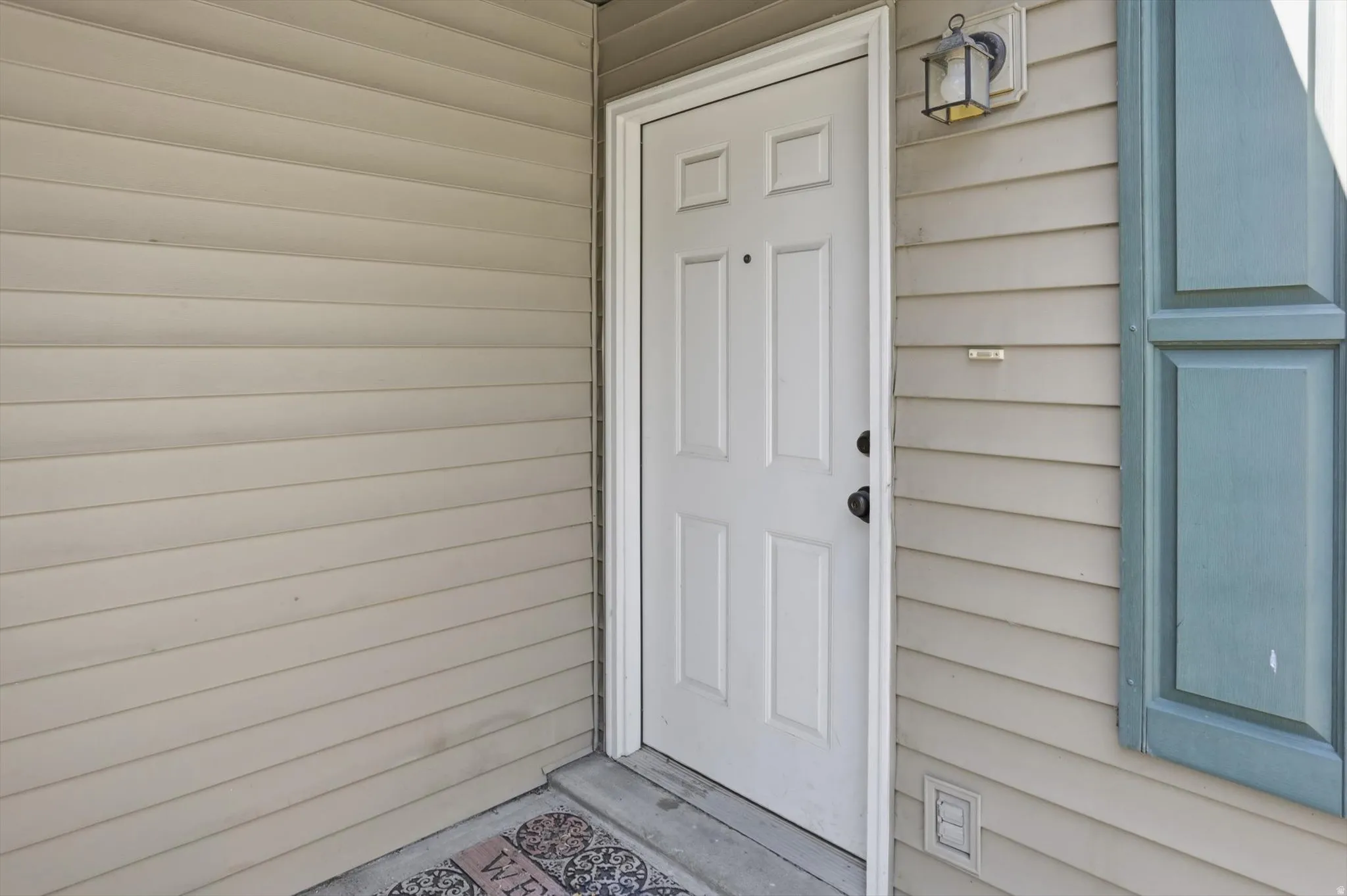 Entry with a four-panel door, tan horizontal siding, and a mounted exterior light fixture