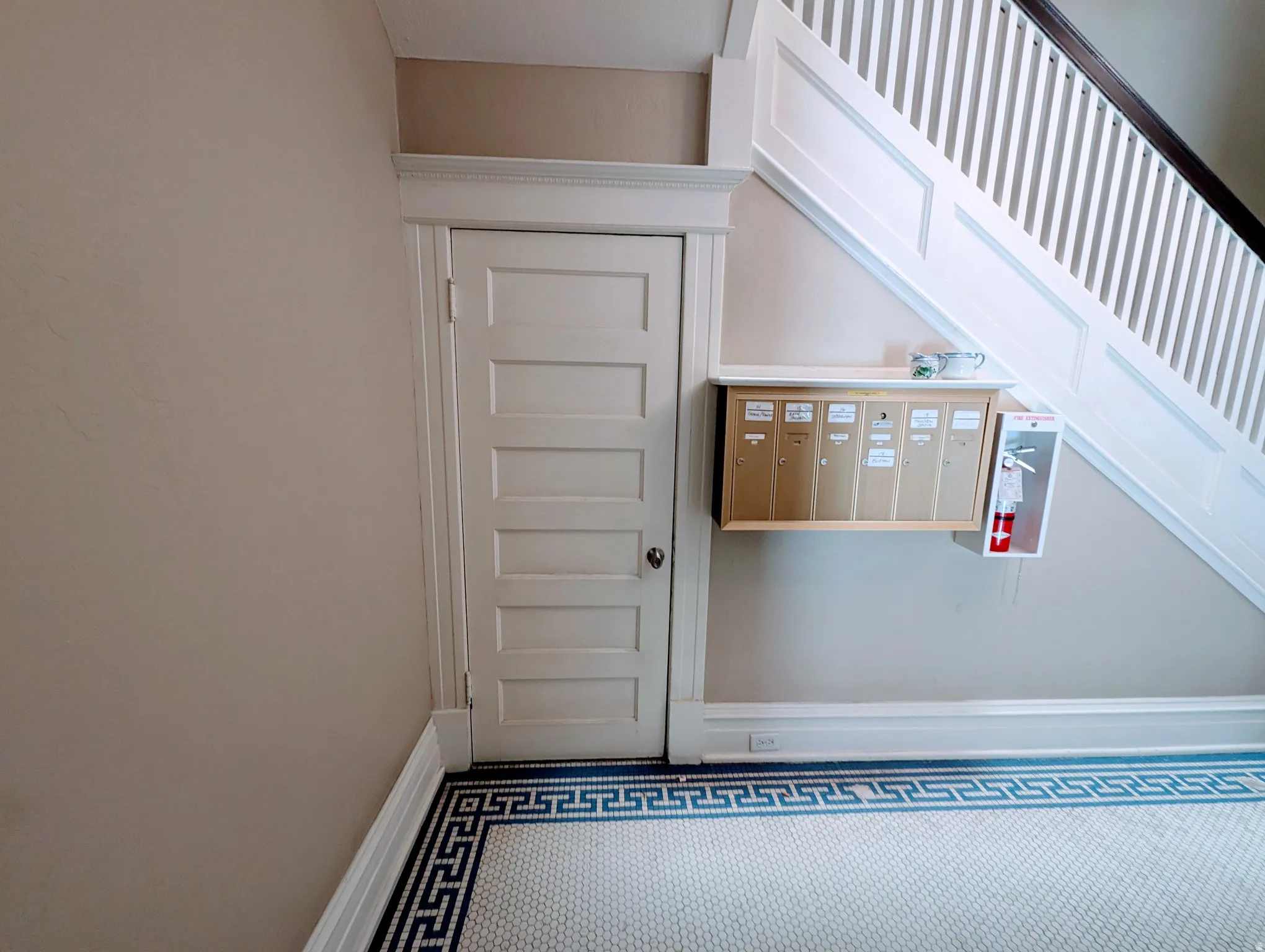 Entryway featuring classic paneled door with detailed trim, a built-in mail station, staircase with white balusters and dark handrail, and mosaic tile flooring with a Greek key border