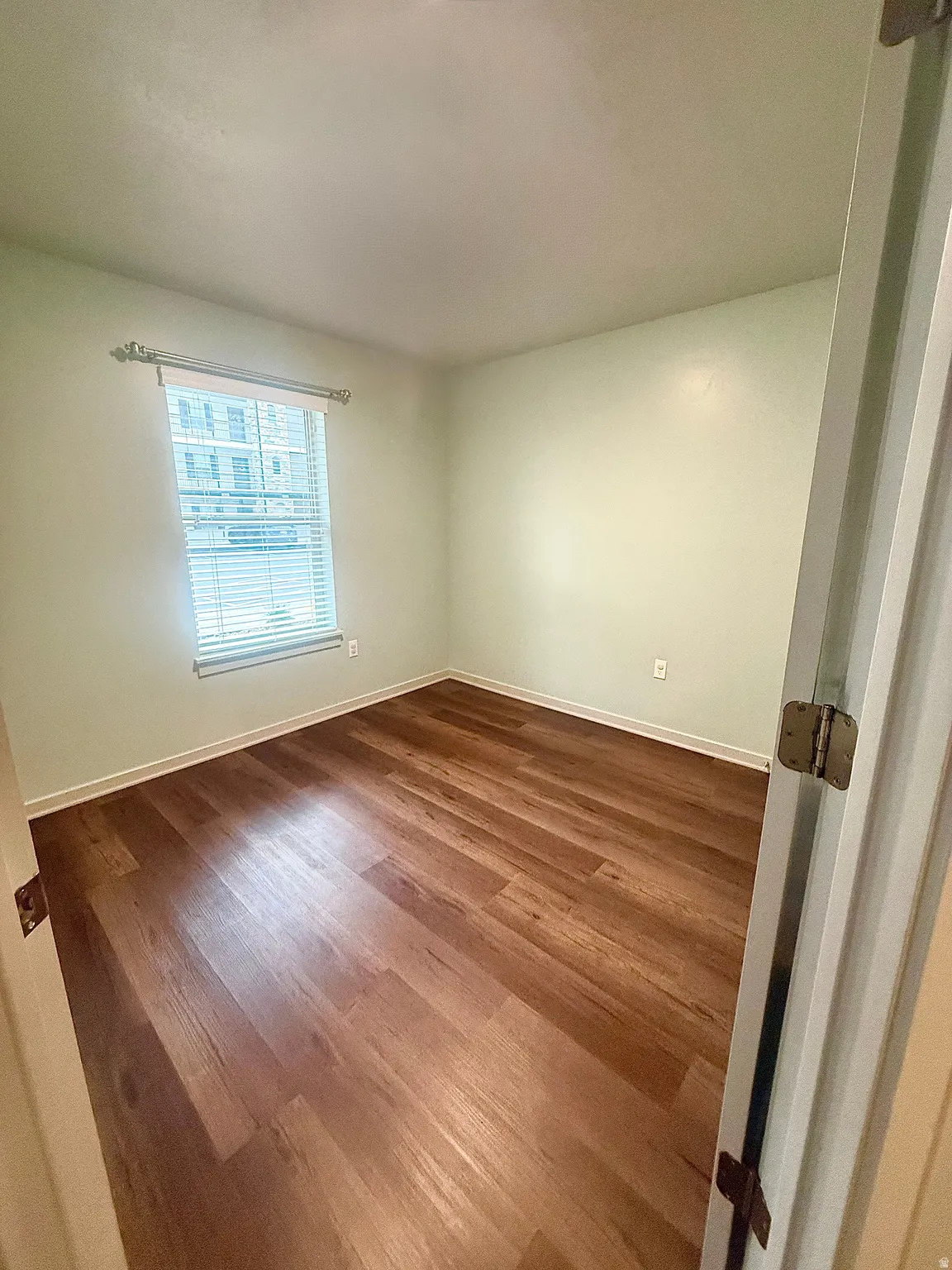 Room featuring wood-finish flooring, light-toned walls, and a window with horizontal blinds
