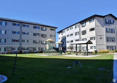 Expansive green space featuring a playground structure and covered picnic area, surrounded by multi-story residential buildings with contrasting siding and dark shutters