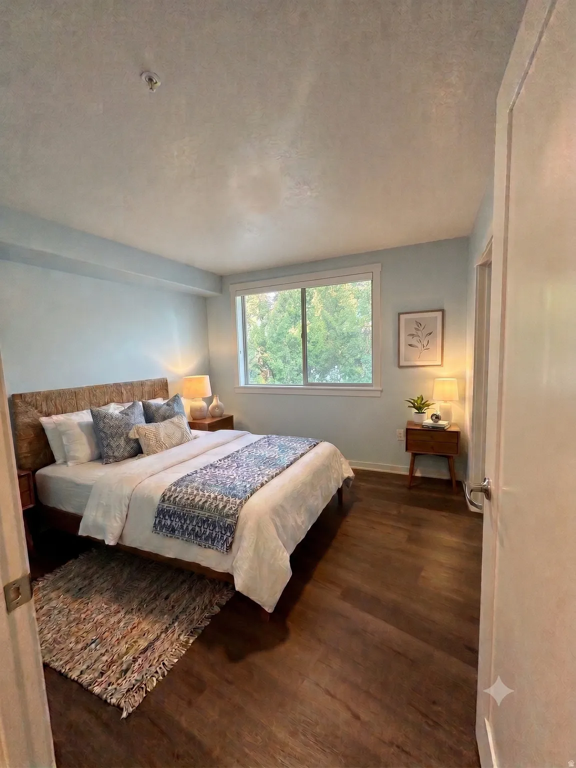 Wood-finish flooring throughout, featuring a broad window with white trim, light blue walls, and a white interior door