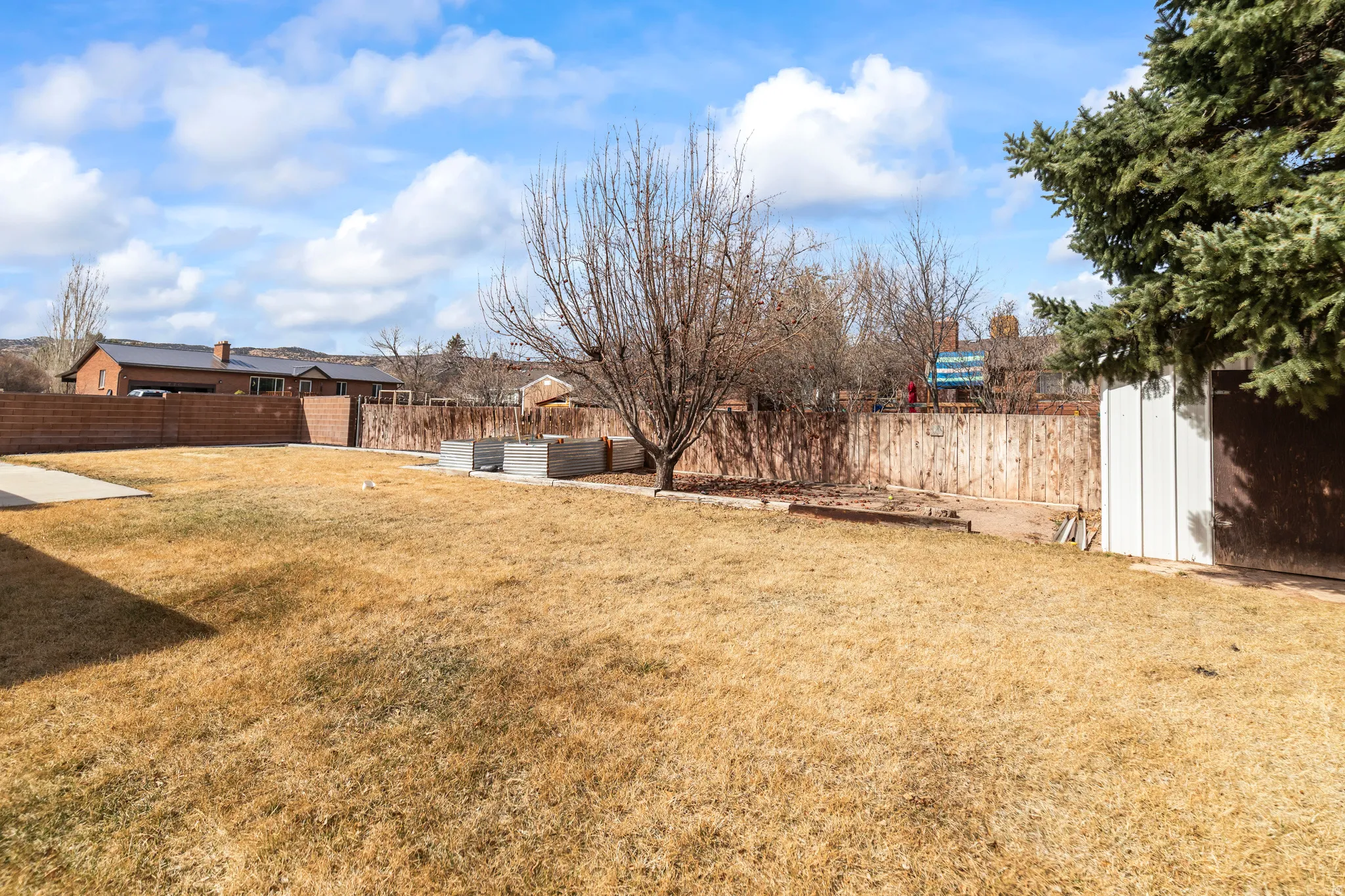Spacious backyard featuring a concrete patio, mature trees, wood fence, brick privacy wall, and a storage shed
