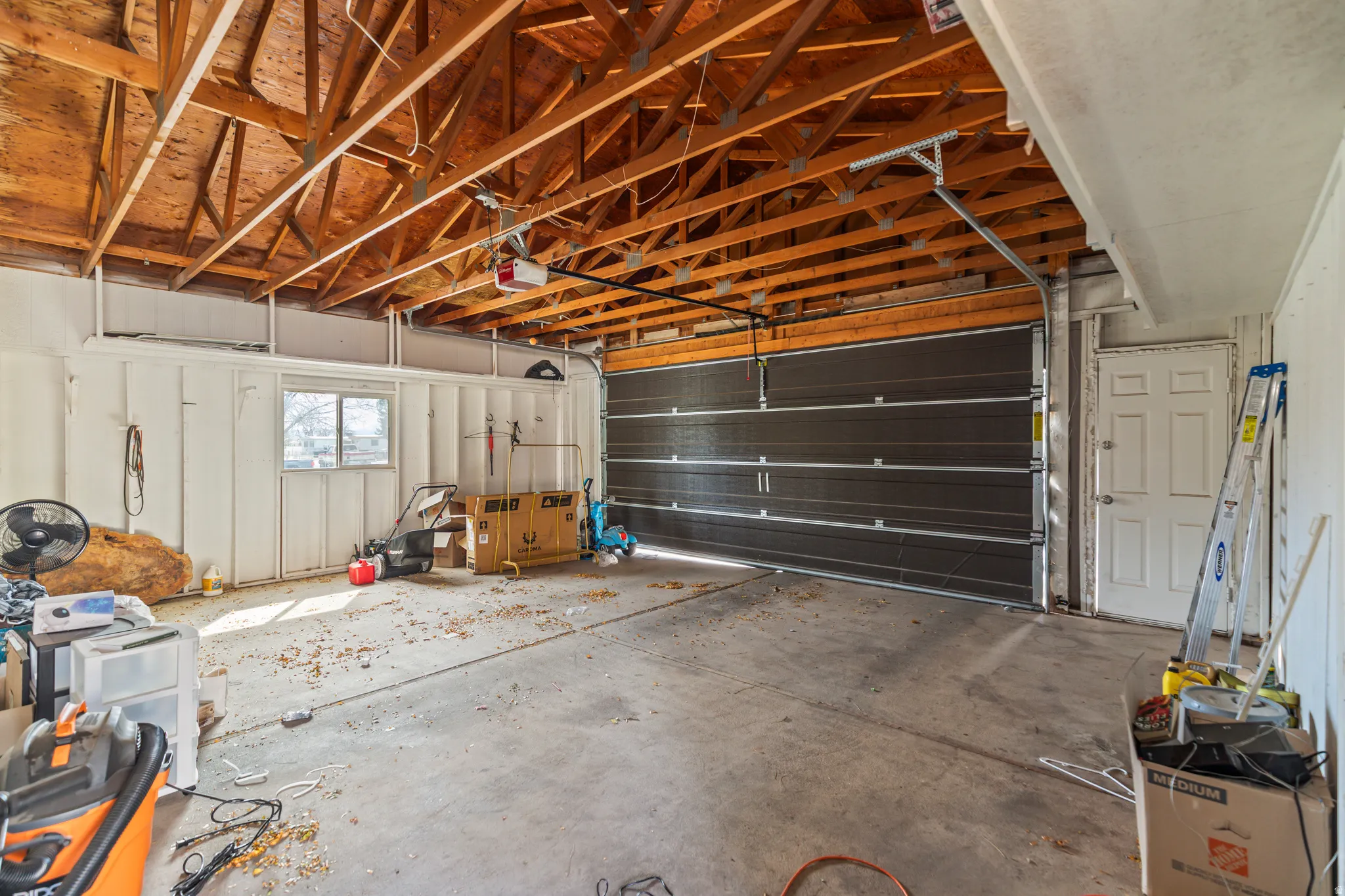 Spacious garage featuring an exposed wood truss ceiling, white paneled walls, a large dark-paneled garage door, a pedestrian door, and a single window