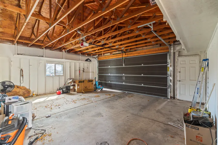 Spacious garage featuring an exposed wood truss ceiling, white paneled walls, a large dark-paneled garage door, a pedestrian door, and a single window
