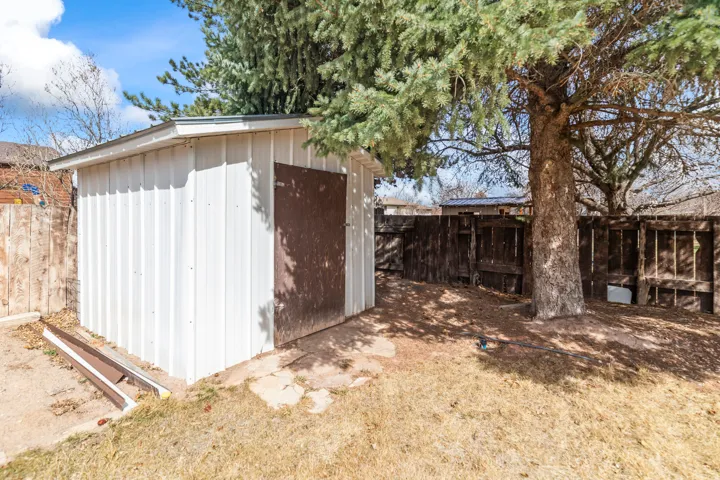 Backyard featuring a white storage shed with a brown door and a sloped roofline, a mature evergreen tree, and a wooden perimeter fence