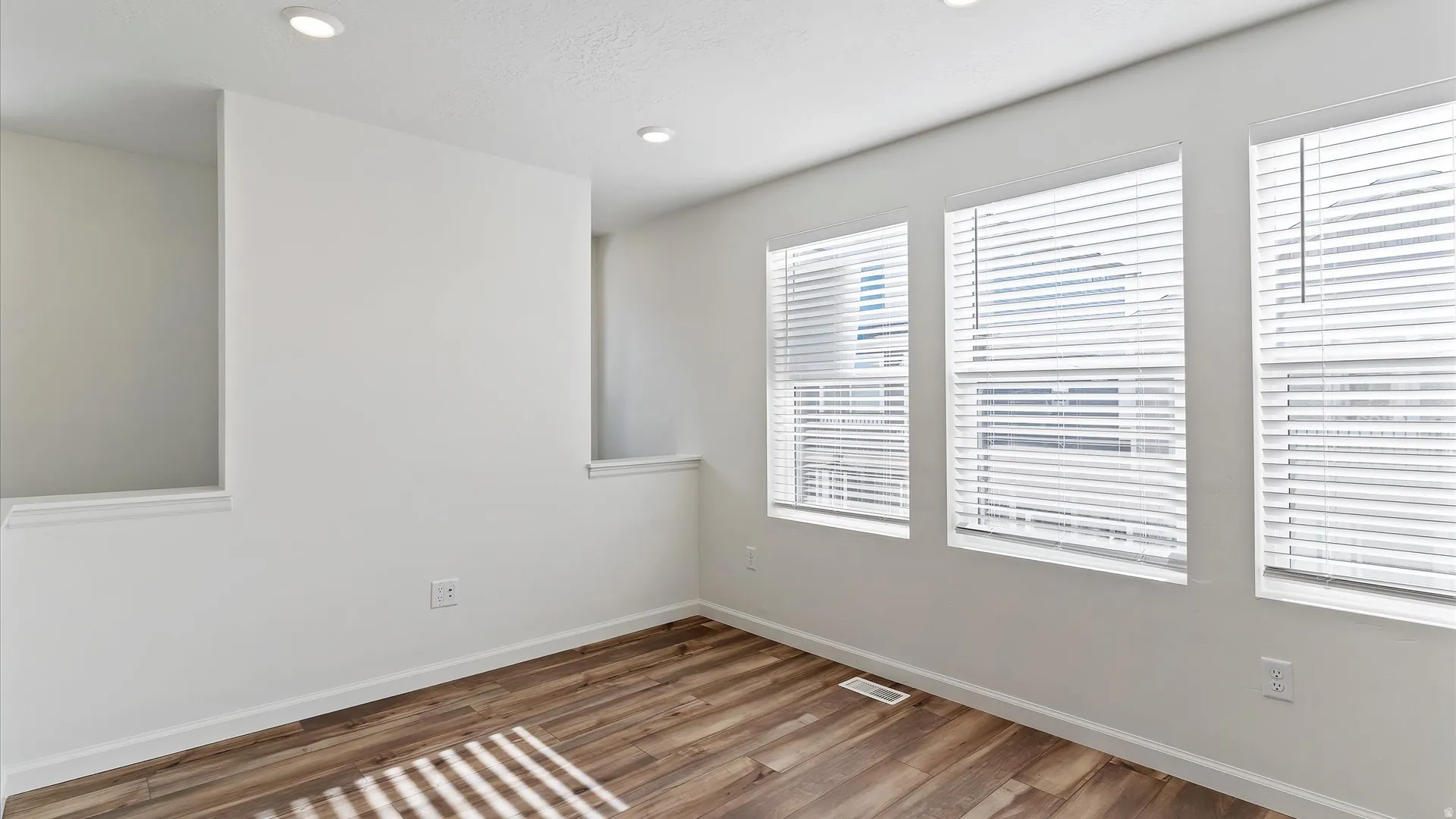 Room featuring wood-finish flooring, three windows with white blinds, recessed lighting, and white walls with built-in wall niches