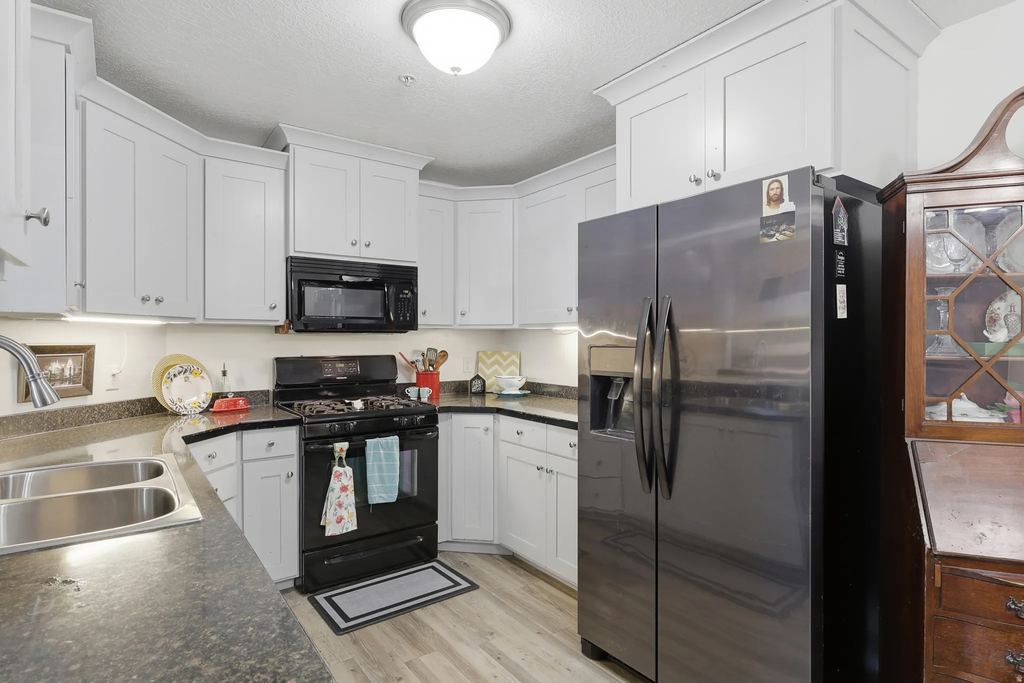 Kitchen featuring L-shaped dark countertops, white cabinetry with crown molding, a stainless steel side-by-side refrigerator, a black gas range, and a dual-basin stainless steel sink