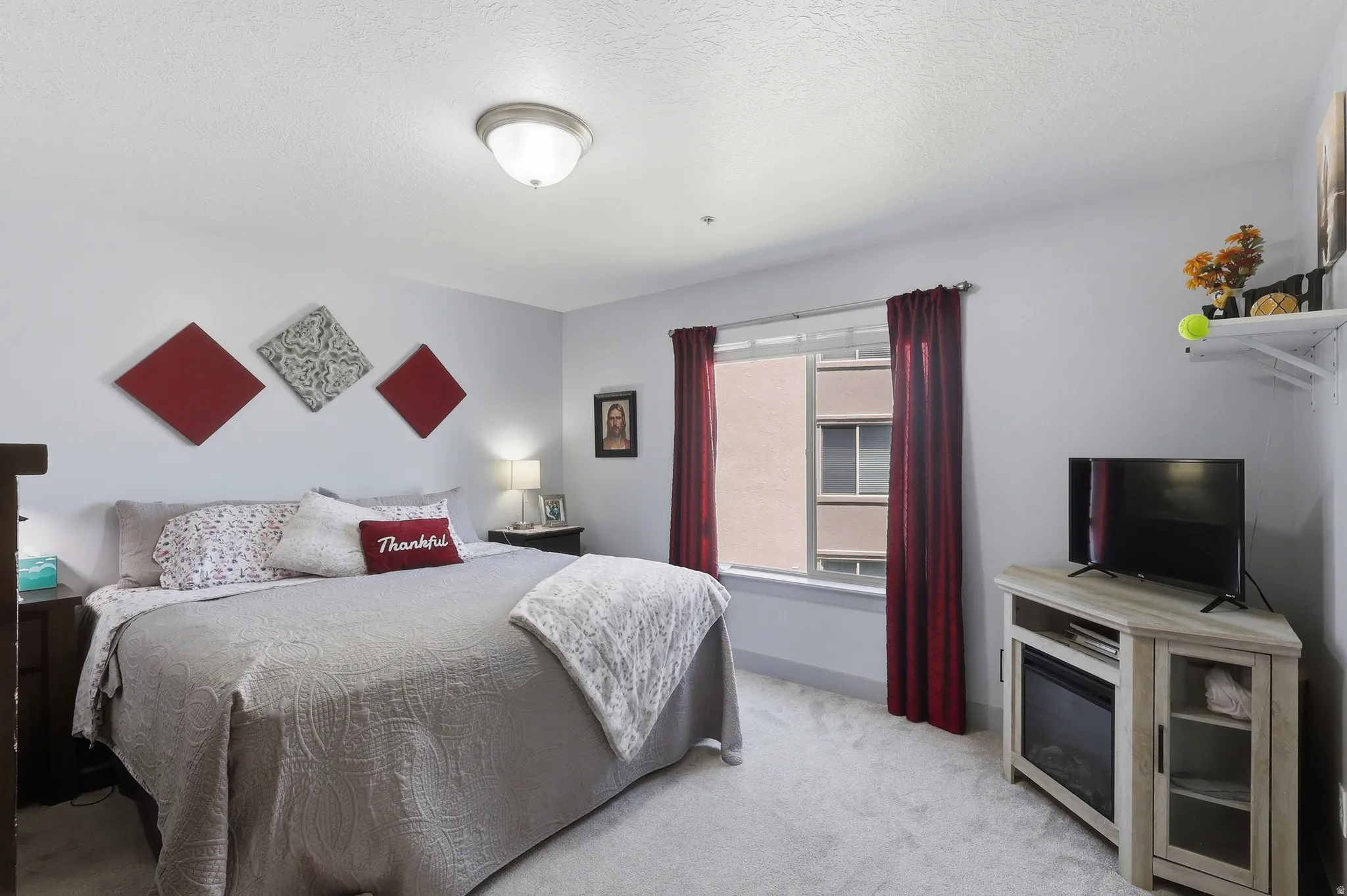 Carpeted bedroom featuring light gray walls, a window with red drapes, and a ceiling-mounted light fixture