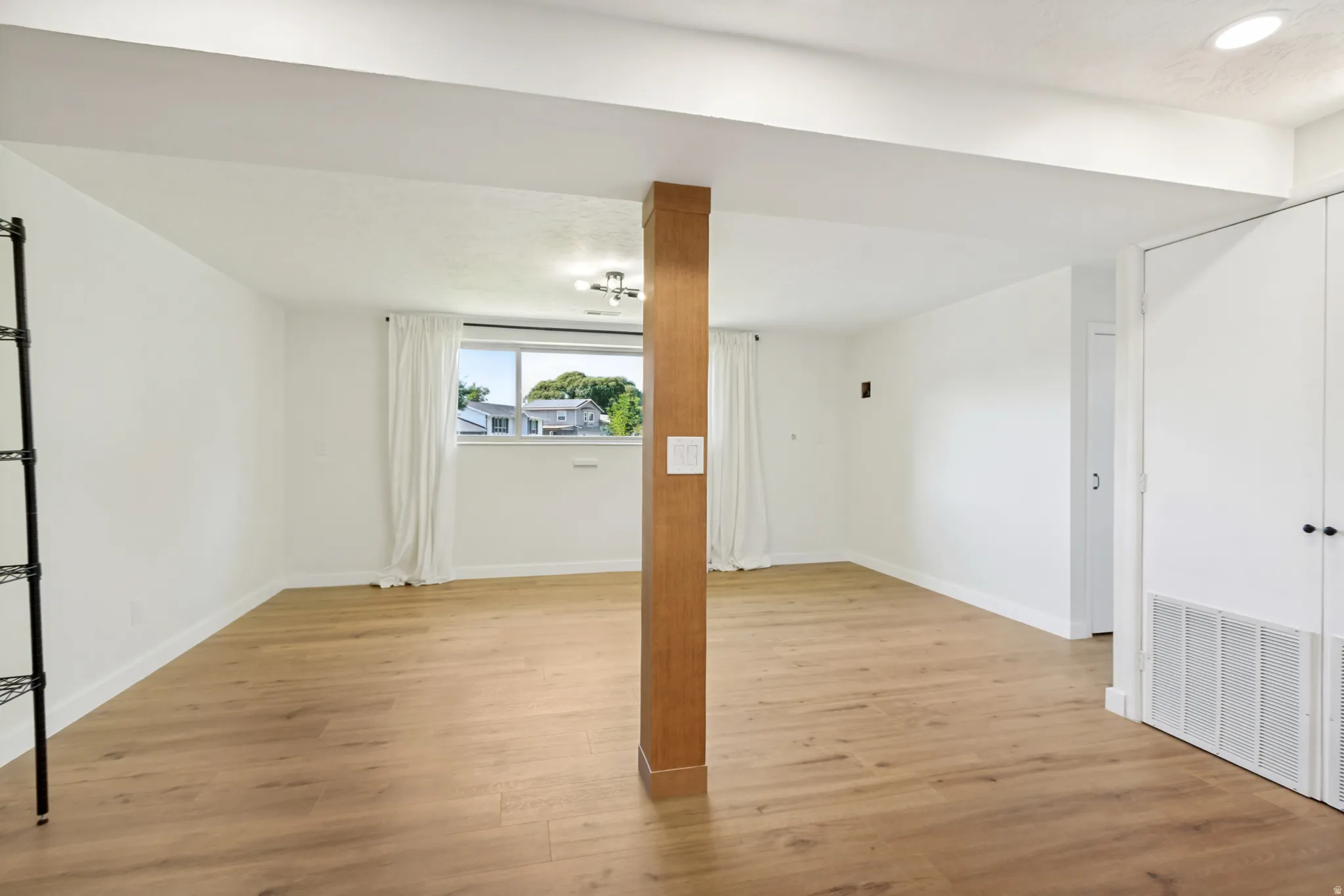 Spacious room featuring light wood-finish flooring, white walls, and a large window with white draperies