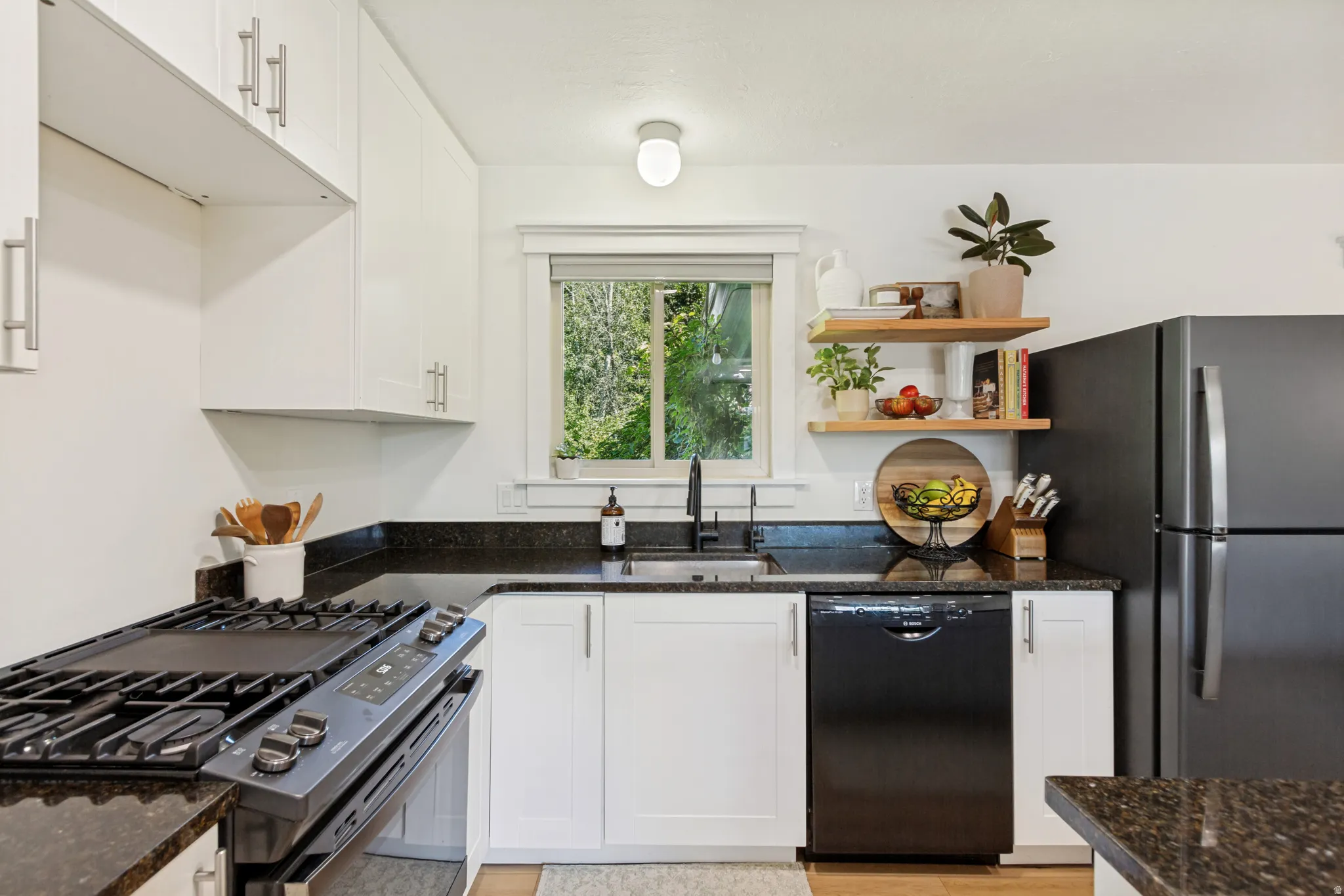 Well-appointed kitchen featuring white cabinetry, dark granite countertops, stainless steel sink with a black faucet, black appliances, and open wood shelving