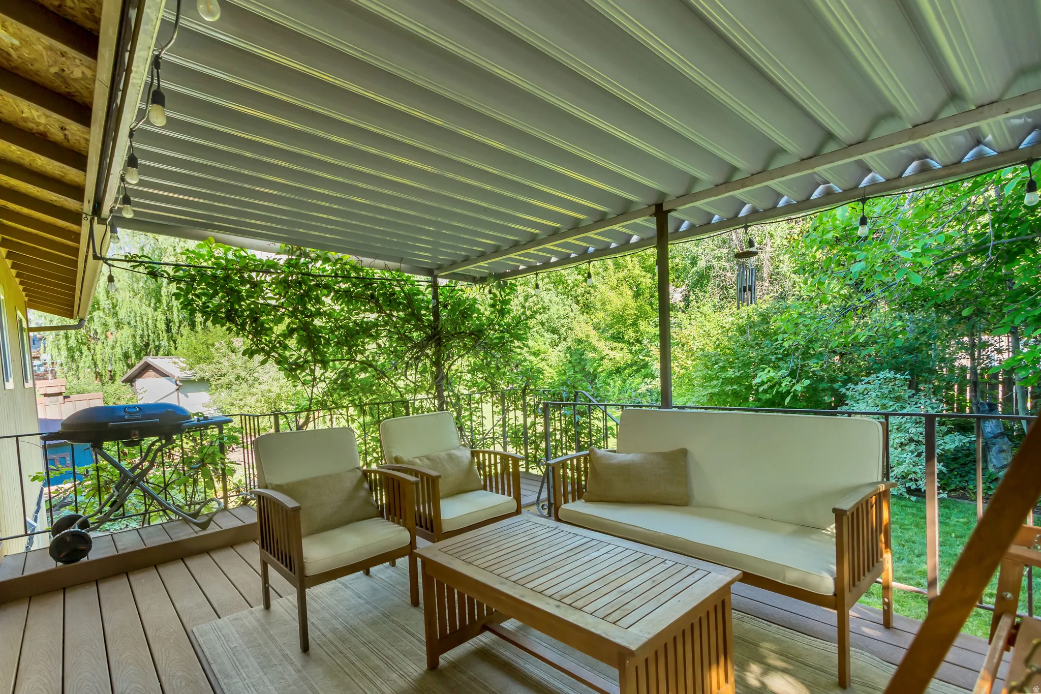 Covered outdoor deck with composite decking, featuring a metal railing, string lighting, and an expansive tree-lined backdrop