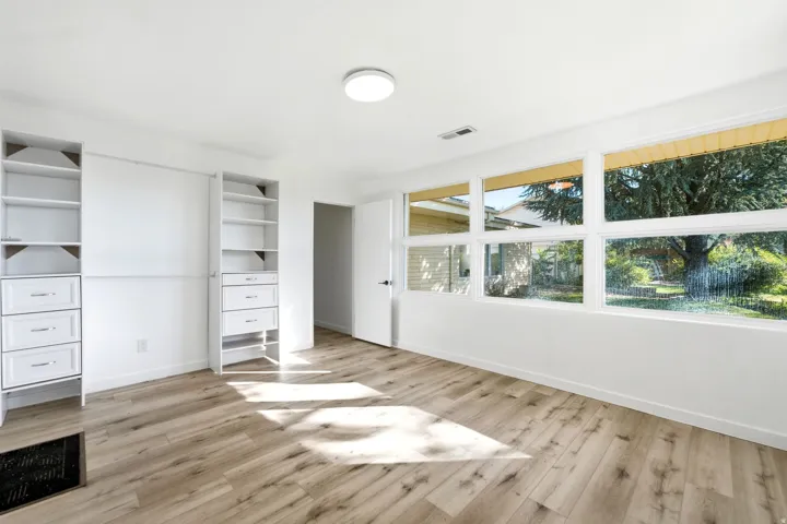 Bright, light-filled room featuring expansive windows with natural outdoor scenery, warm wood-finish flooring throughout, and built-in cabinetry with shelving along the left wall