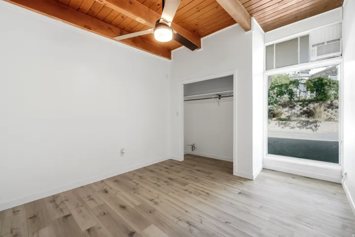 Bright corner bedroom with wood-finish flooring, exposed beam ceiling with recessed lighting, large windows with natural light, and a spacious closet with sliding door