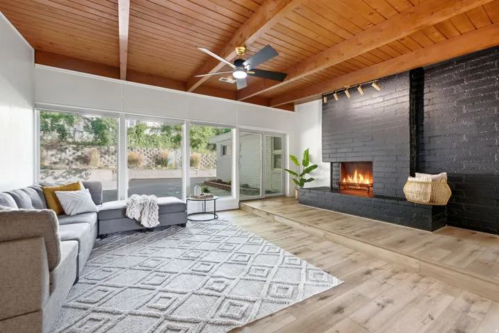 Vaulted wood-beamed ceiling, floor-to-ceiling windows, black brick fireplace with an elevated hearth, wood-finish flooring, and a ceiling fan