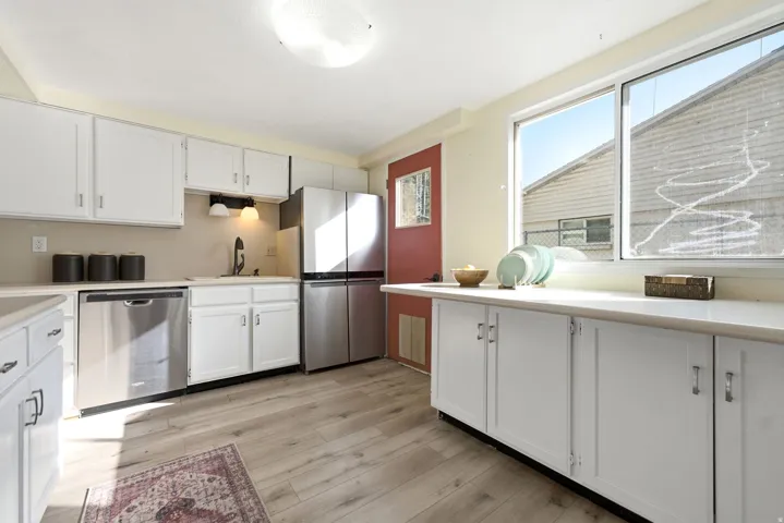Bright, updated kitchen featuring white cabinetry, stainless steel appliances including a dishwasher and refrigerator, wood-finish flooring, and abundant natural light from large windows
