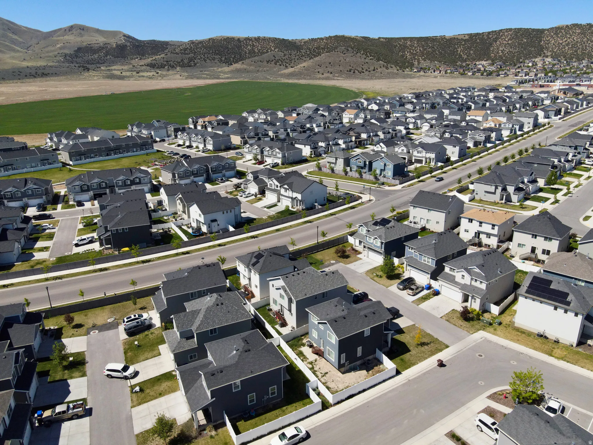 Expansive residential development featuring numerous single-family homes and townhomes, surrounded by a large green field and distant mountains