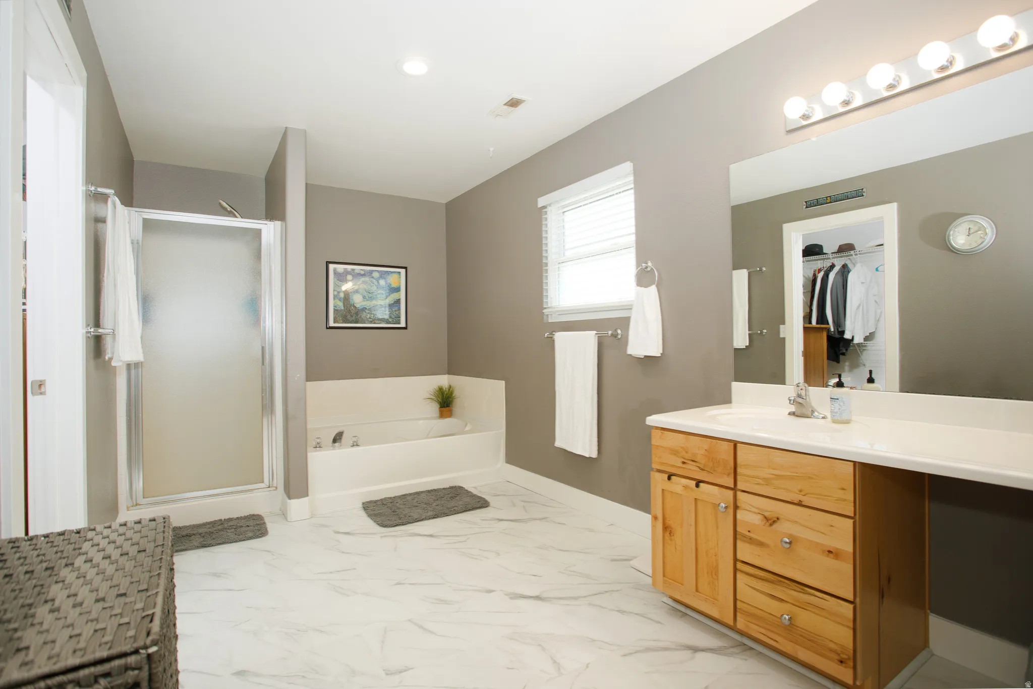 Spacious bathroom featuring large format tile flooring, a glass-enclosed shower, a built-in soaking tub, a single vanity with a light wood-finish cabinet, and a large mirror