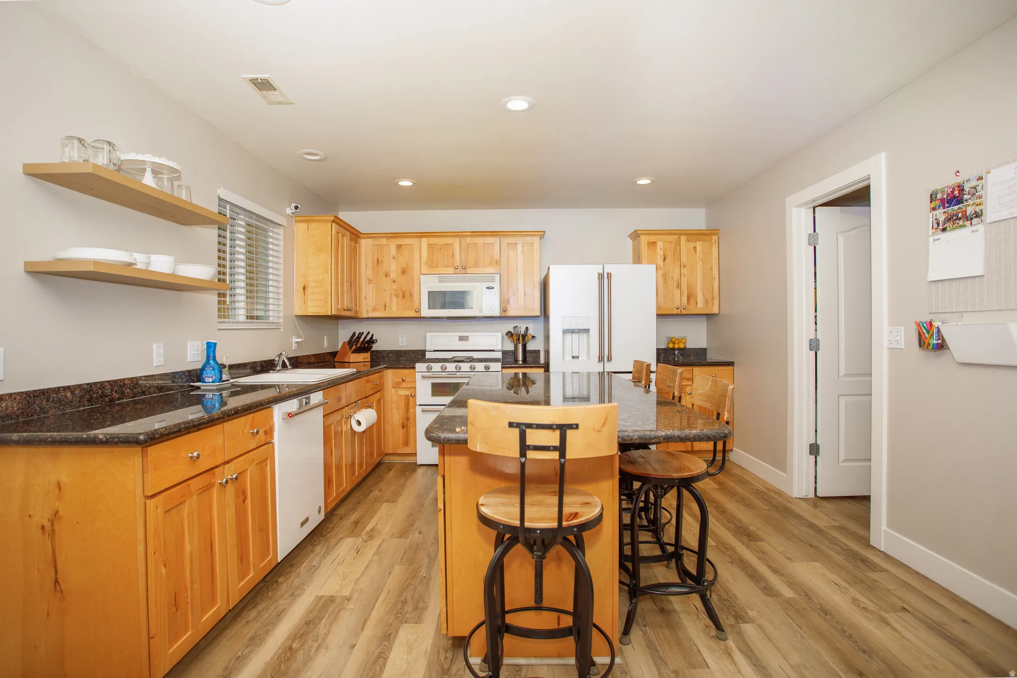 Kitchen featuring wood-finish flooring, a central island with countertop seating, wood cabinetry, dark stone-finish countertops, and white appliances
