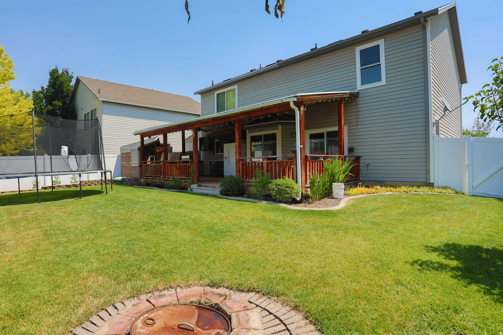 Spacious backyard featuring a covered patio, expansive lawn, mature landscaping, and a paver-lined fire pit