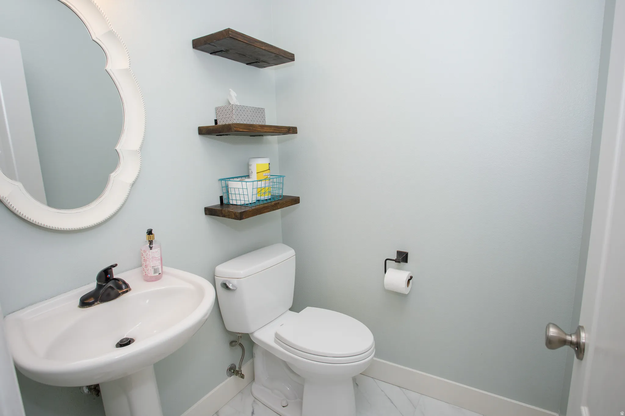 Pedestal sink with dark bronze faucet, white scalloped mirror, and three wall-mounted wood-finish shelves