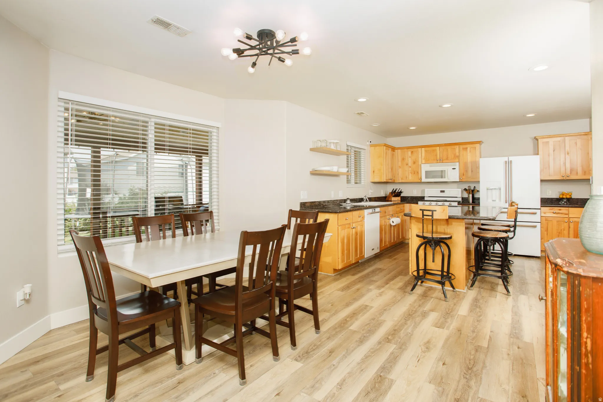 Open-concept kitchen and dining area featuring wood-finish flooring, a contemporary ceiling light fixture, and a large window with blinds