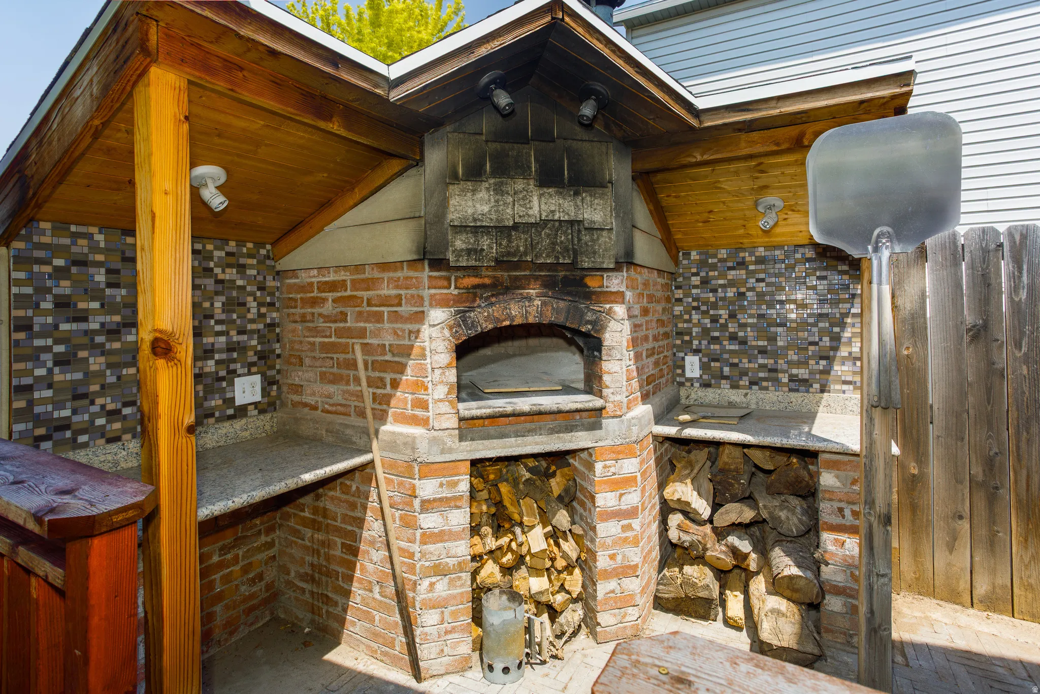 Outdoor brick oven with wood storage, built-in countertops, and mosaic tile backsplash under a wood-paneled covered structure