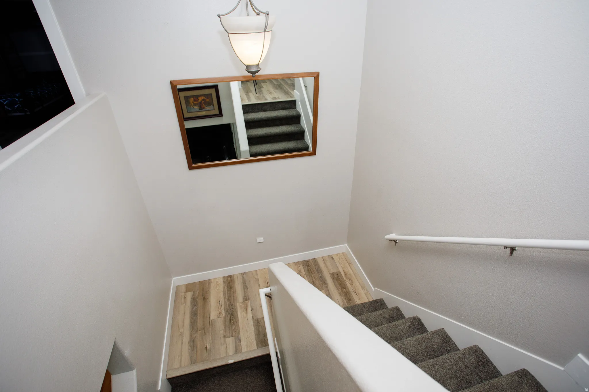 Staircase featuring wood-finish flooring, soft gray carpeting, a white handrail, a rectangular framed mirror, and an overhead pendant light fixture