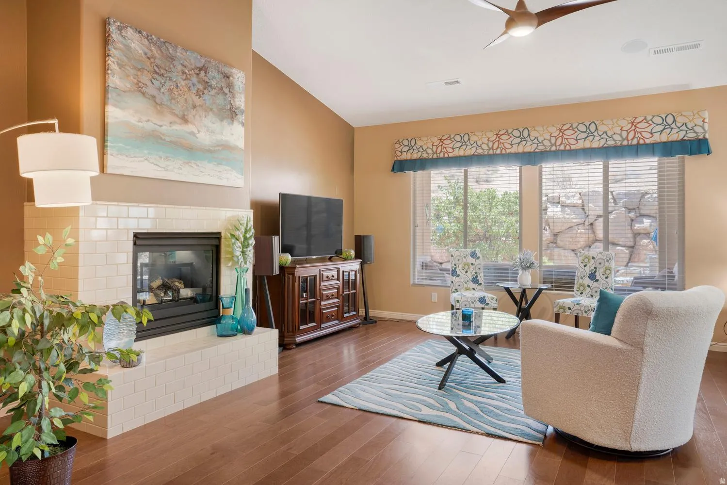 Spacious living area featuring wood-finish flooring, a tiled fireplace with a stone surround, and large windows with blinds and valances
